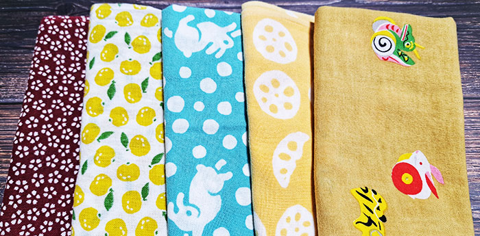 異なるイラストや柄が名入れ印刷された5種類の異なる手ぬぐいが折りたたんだ状態で並んでいる画像