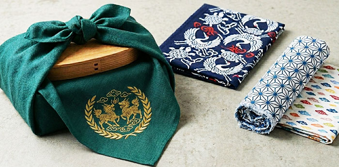 木製の箱を包んだ緑色の風呂敷と絵柄が異なる2枚の折りたたまれた手ぬぐいと、筒状に丸められた手ぬぐいが一緒に写っている画像
